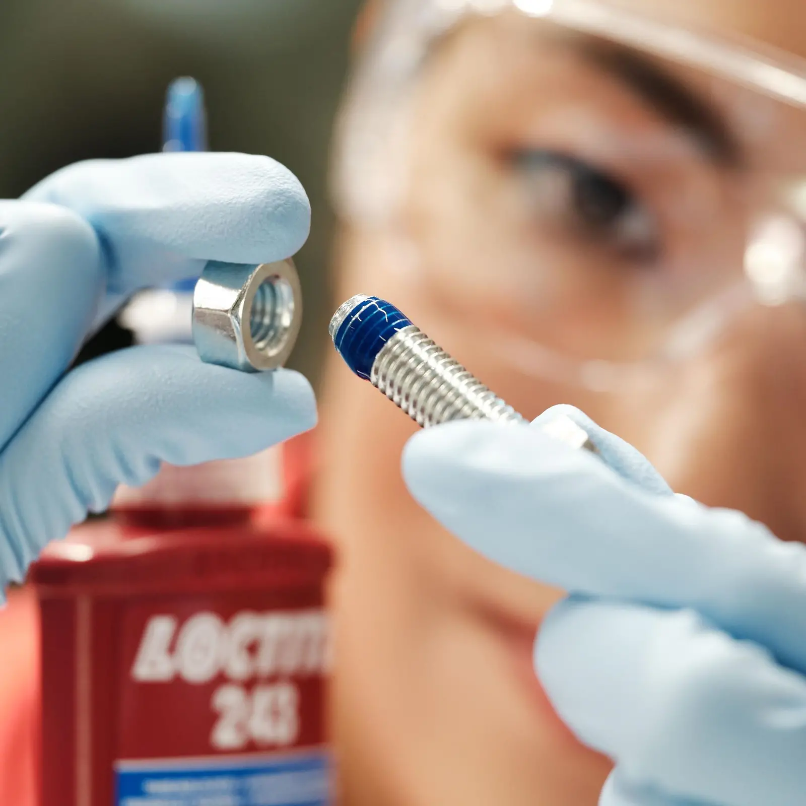 Het gezicht van een vrouw op de achtergrond. Op de voorgrond haar handen met blauwe handschoenen die een schroef met blauwe Threadlocker vasthouden.