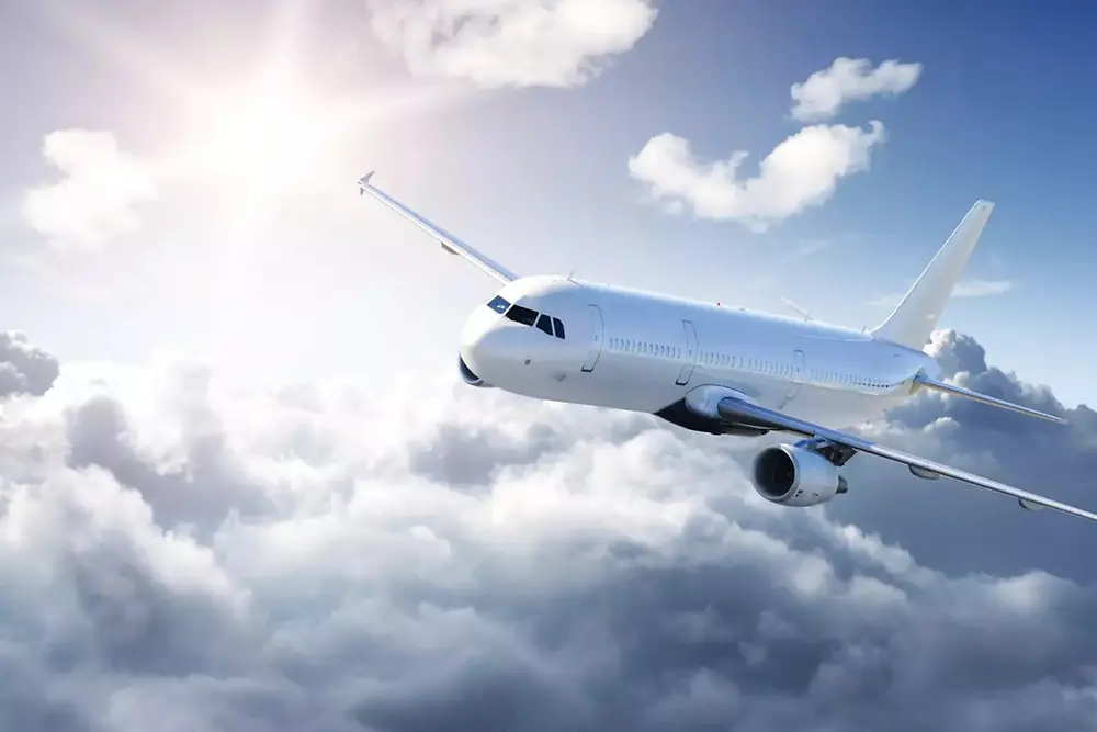 Un avion de ligne blanc vole au-dessus de nuages doux dans un ciel lumineux, avec la lumière du soleil venant de la gauche en haut.