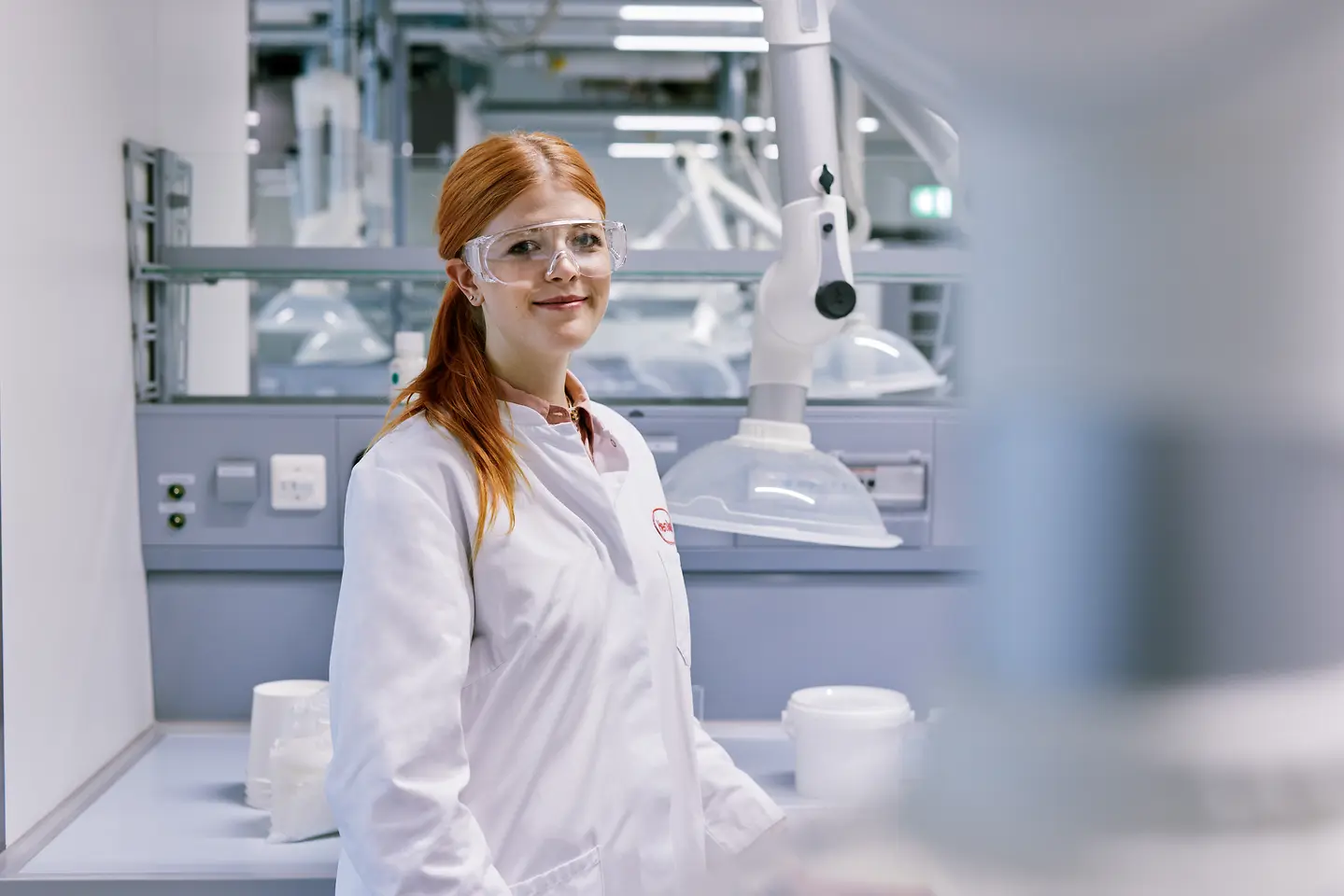 Un jeune diplômé débutant sa carrière chez Henkel, vêtu d'une blouse blanche de laboratoire, debout dans un laboratoire moderne avec du matériel technique en arrière-plan.