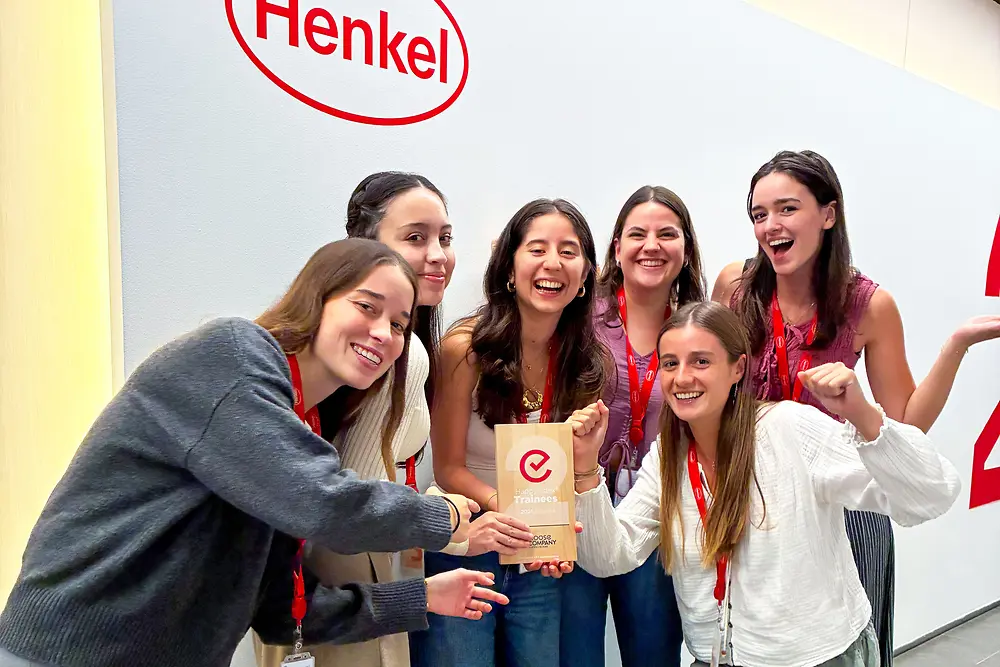 Un groupe de six personnes se tient debout devant un mur avec le logo Henkel. Elles portent des tours de cou rouges et des tenues décontractées, et sont rassemblées autour d'une plaque de récompense. Le cadre semble être un espace d'entreprise intérieur avec un mur de couleur claire et un sol gris. Le groupe pose avec enthousiasme, mettant en valeur le prix au centre.