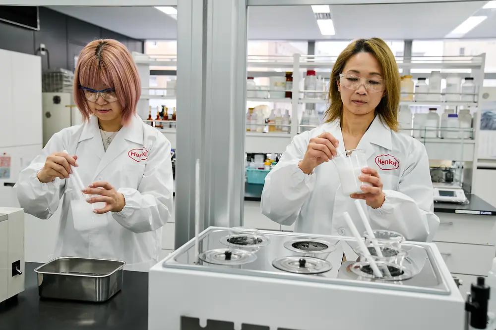 Deux personnes portant des blouses de laboratoire Henkel blanches travaillent dans un laboratoire moderne. Elles manipulent des instruments scientifiques et des récipients sur un plan de travail, avec divers équipements de laboratoire et des étagères de fournitures visibles en arrière-plan.