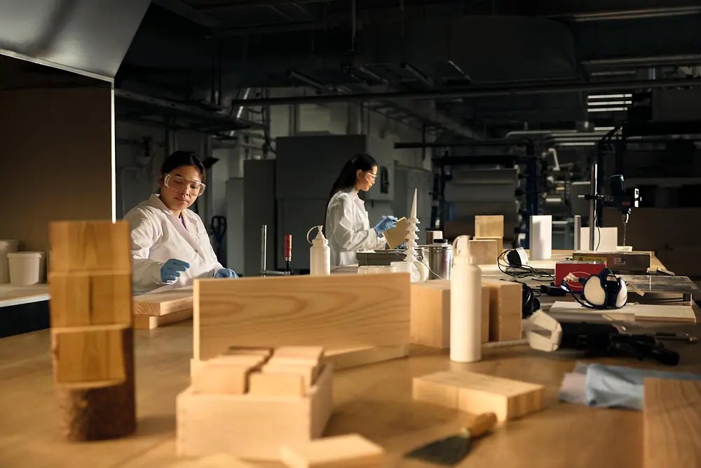 Atelier collaboratif réunissant des personnes portant des blouses et des gants de laboratoire, travaillant autour d’une grande table en bois recouverte d’échantillons de bois, d’outils et de flacons.