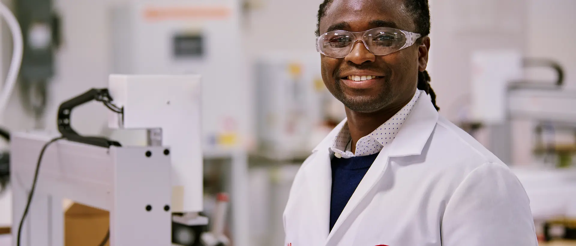 Een persoon in een witte labojas van Henkel in een laboratorium, omgeven door apparatuur en machines.