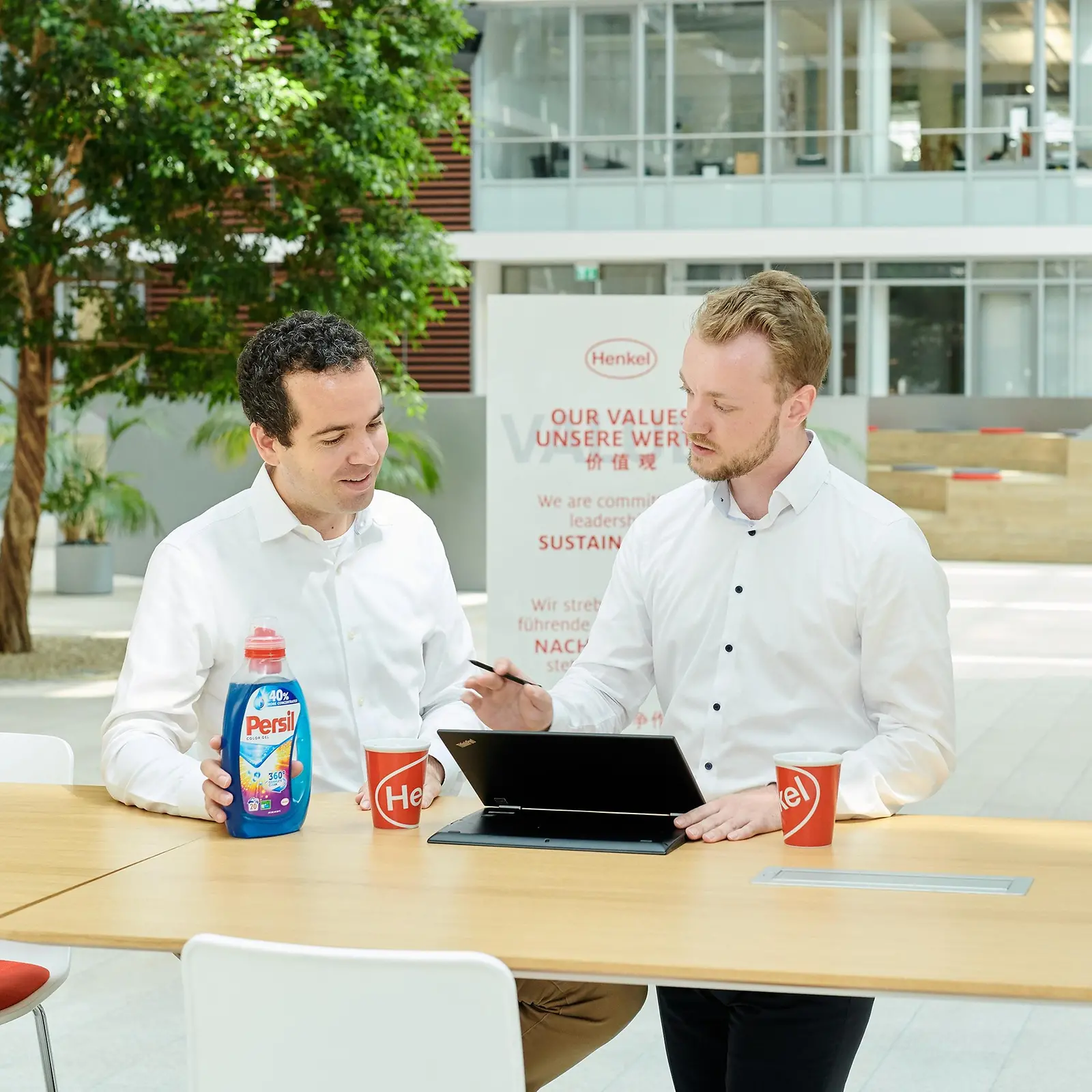 Twee mannen aan een tafel met een fles Persil en een laptop